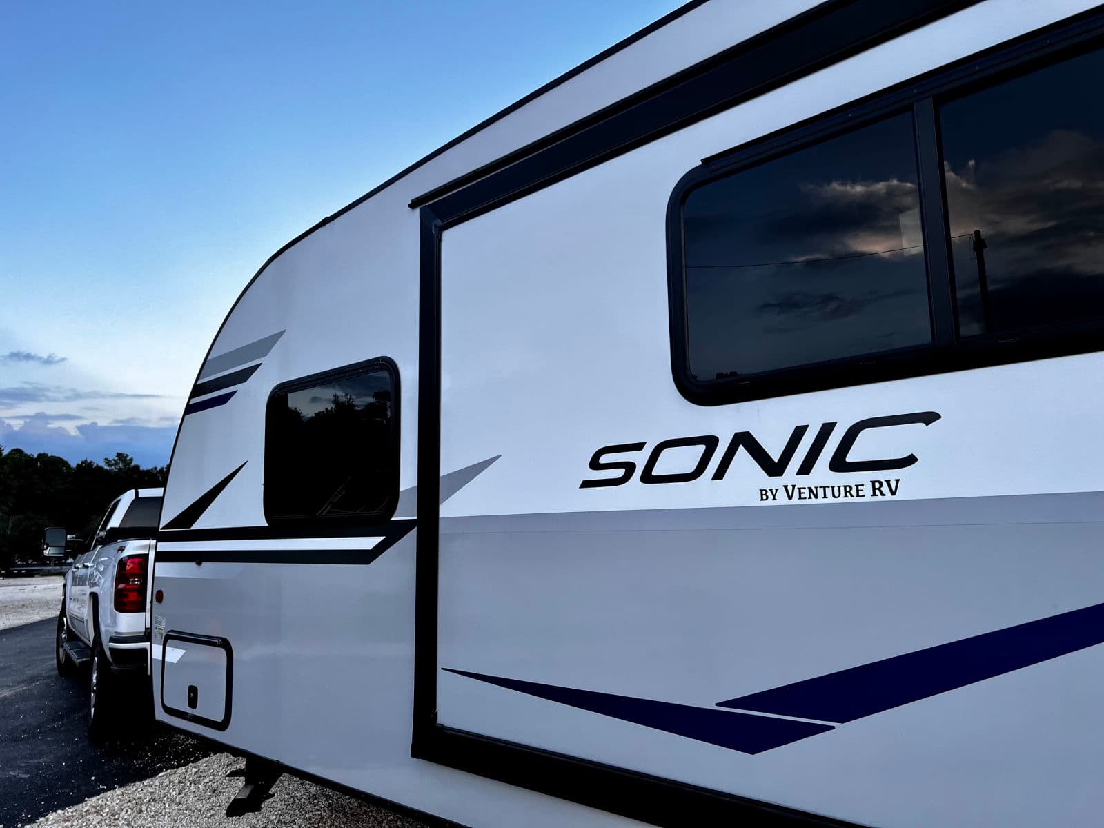 Modern white Sonic RV by Venture RV, hooked to a pickup truck, ready for an RV rental adventure under a cloudy sky.