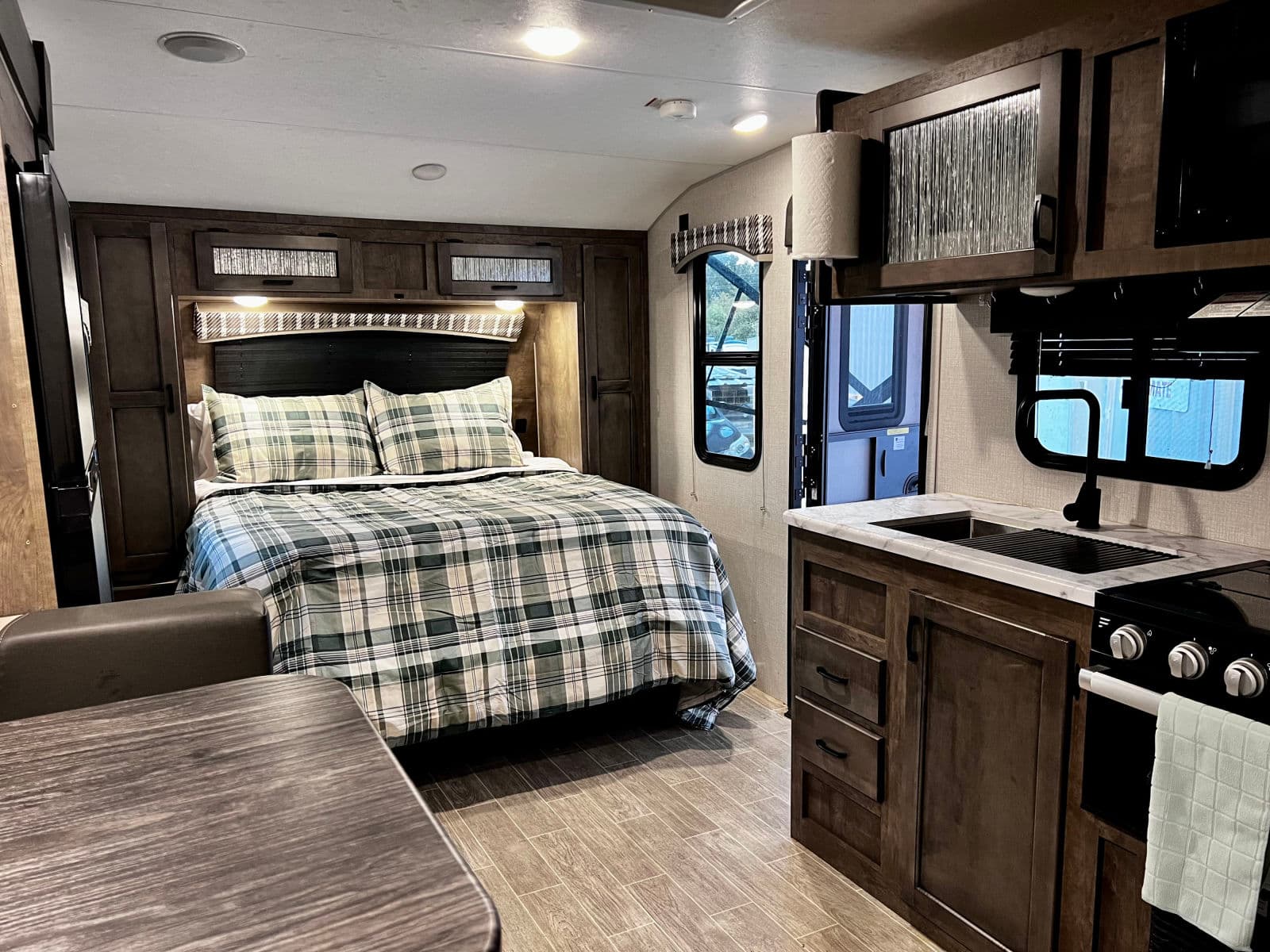 Experience comfort in this modern RV rental. Features a cozy bedroom with plaid bedding and a compact kitchen with sink and stove.