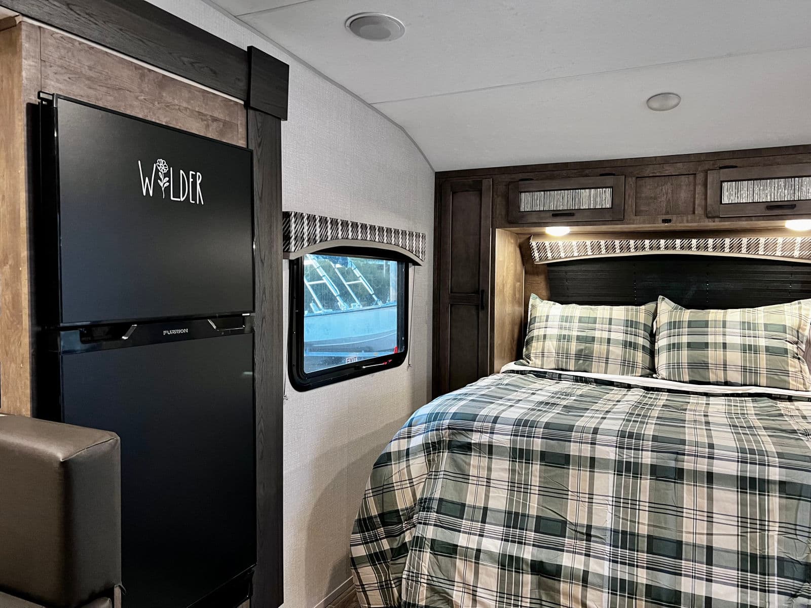Discover comfort in this RV rental's cozy bedroom, showcasing a plaid bed, window, Furrion fridge, and overhead cabinets.