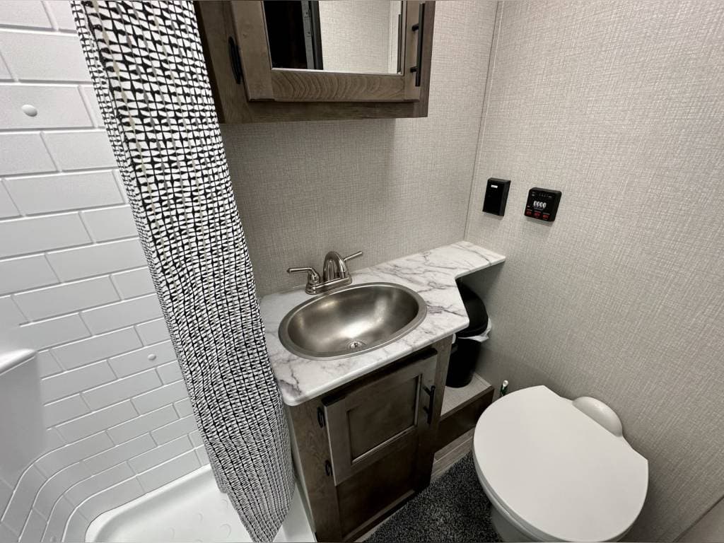 RV rental's compact bathroom features a shower with patterned curtain, sink, wood vanity, toilet, and wall controls.