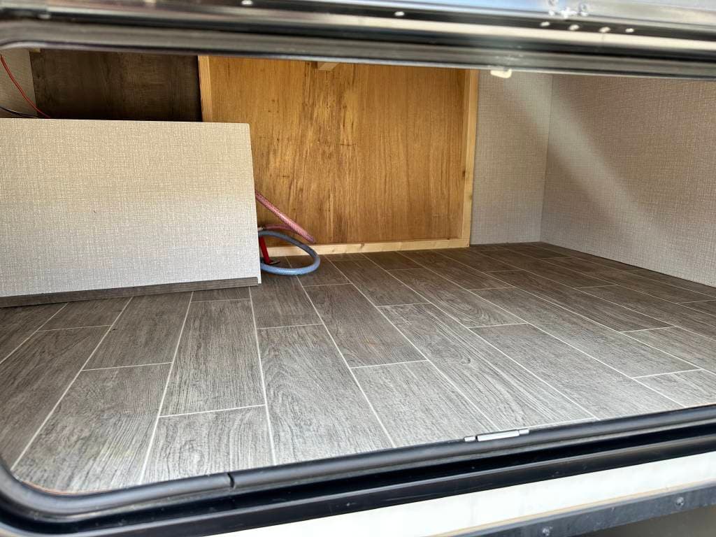Empty RV rental storage compartment with wood-look floor. Hoses visible at the back for utilities.