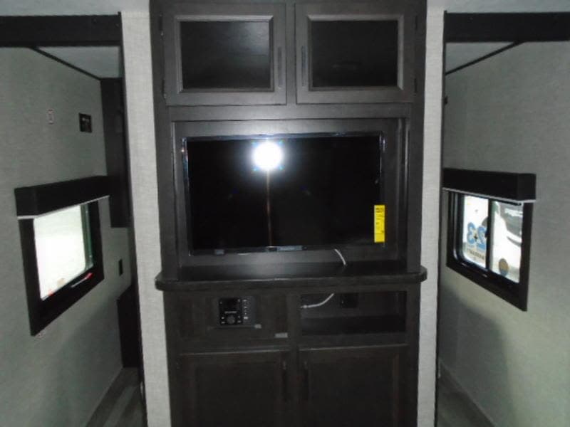 RV rental interior with a dark entertainment unit, television, and cabinets, flanked by narrow passages and windows.