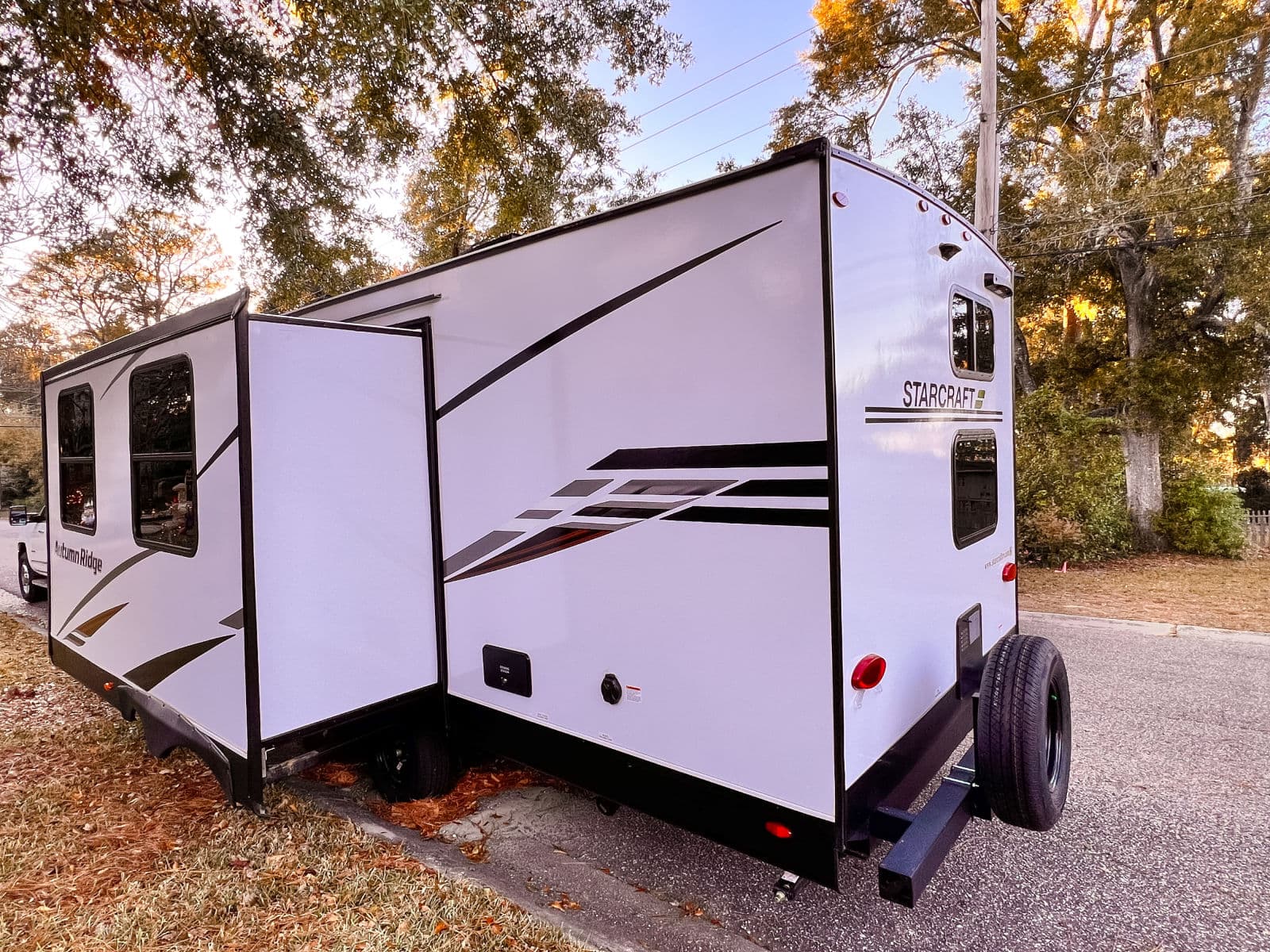 Exterior view of a white Starcraft Autumn Ridge RV rental with its slide-out extended, parked by a road with trees overhead.