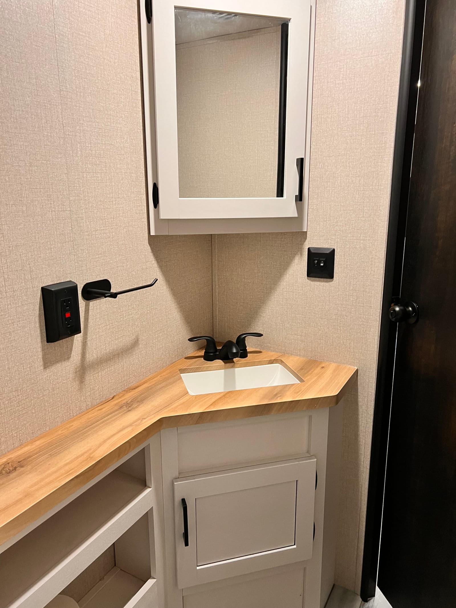 Inside an RV rental bathroom, featuring a corner sink, wooden countertop, mirrored medicine cabinet, and black hardware.
