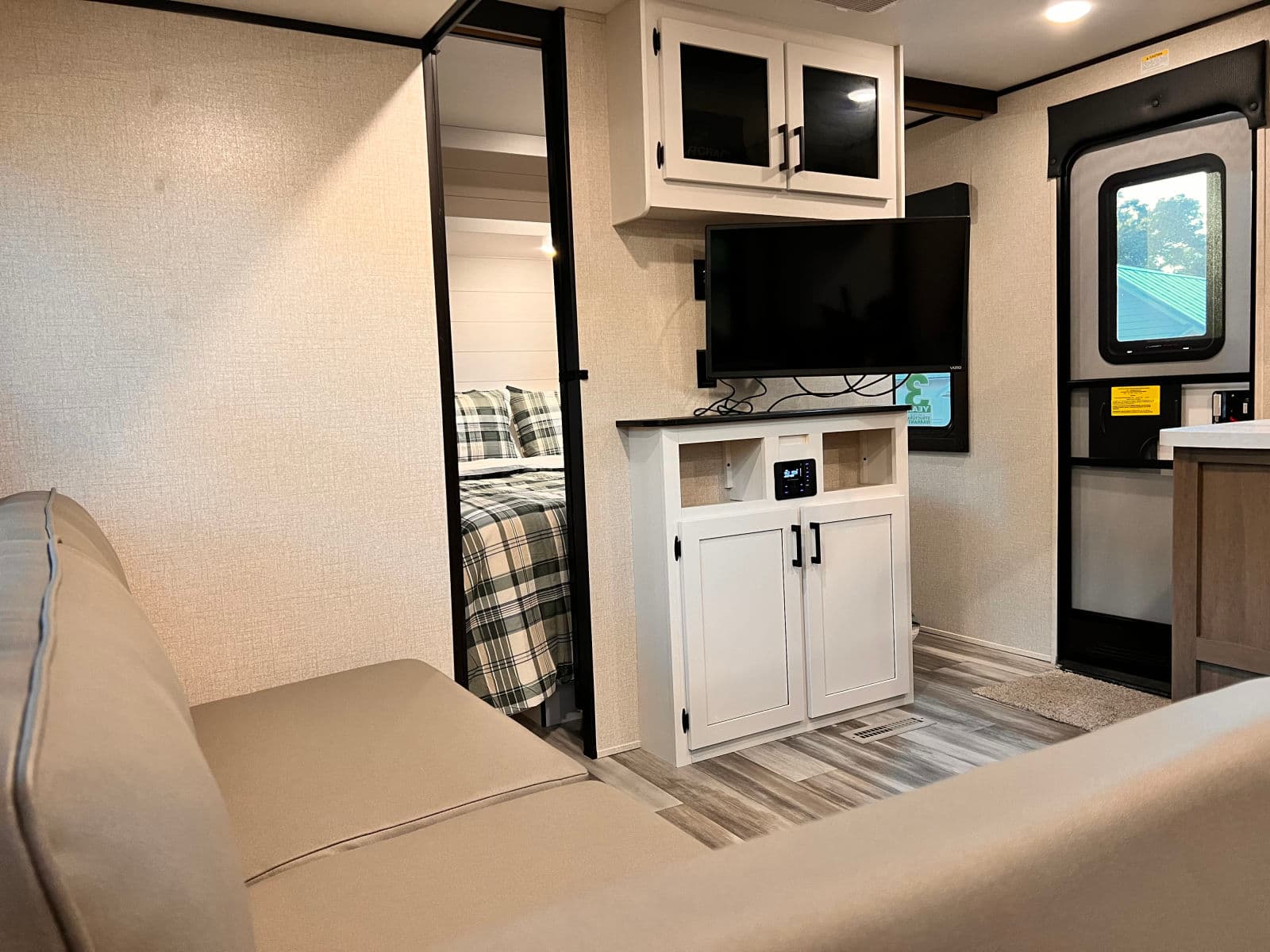Cozy RV rental living area featuring a light sofa, entertainment center with TV, and a glimpse into a bedroom with plaid bedding.