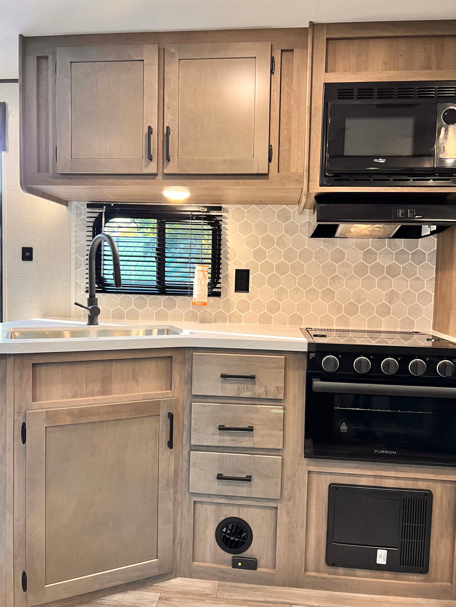 RV rental kitchen: light wood cabinets, black faucet, stove, microwave, and a light honeycomb backsplash.