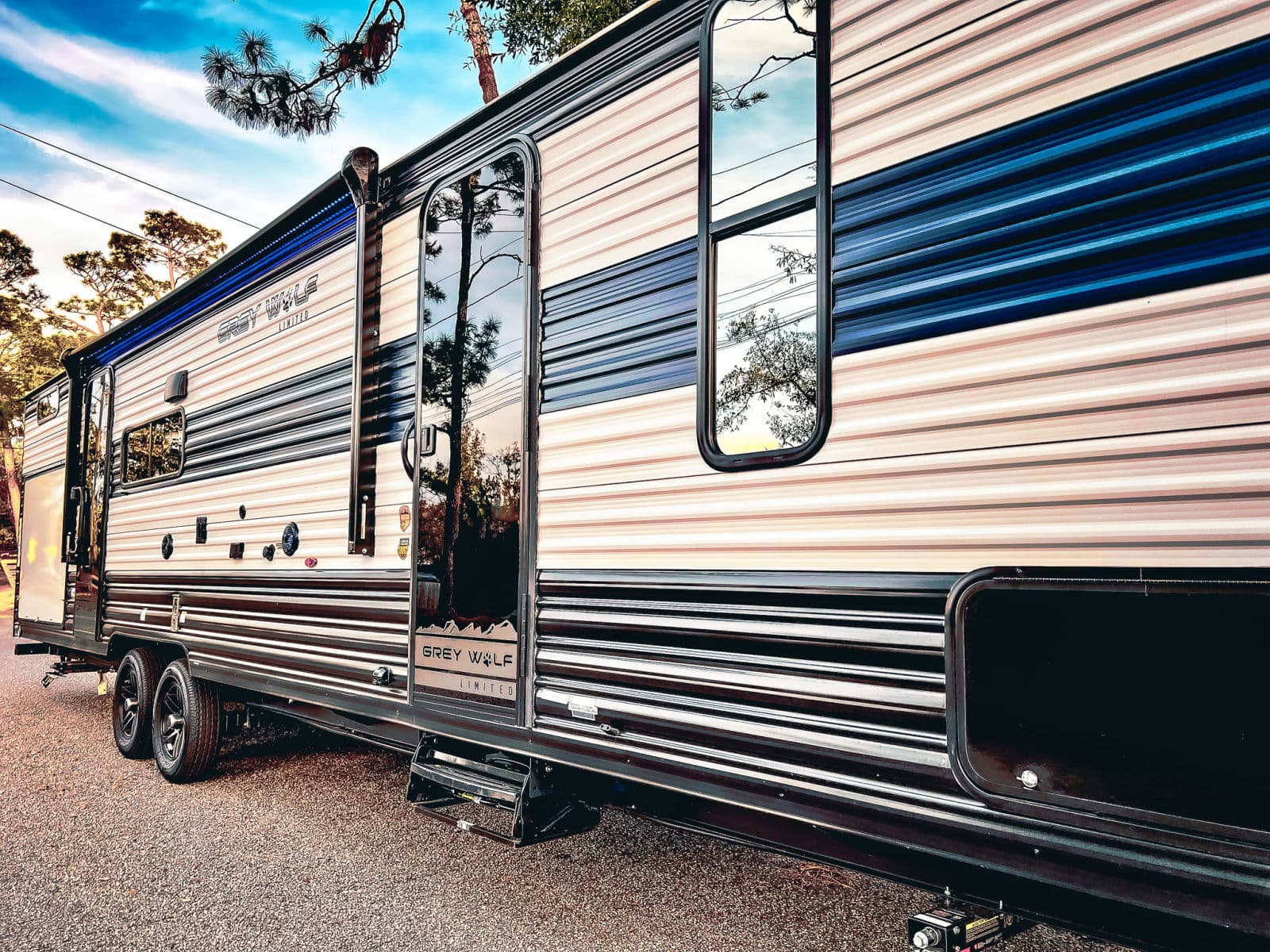 This stylish Grey Wolf Limited RV rental, parked on gravel, features blue LED awning lights and window reflections of trees and sky.