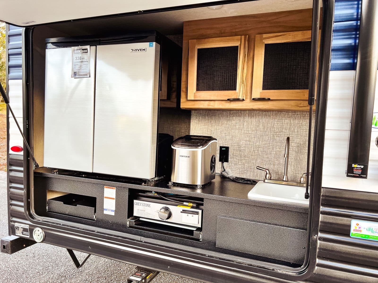 This rv rental outdoor kitchen features a stainless fridge, sink, Greystone pull-out grill, Contoure ice maker, and cabinets.