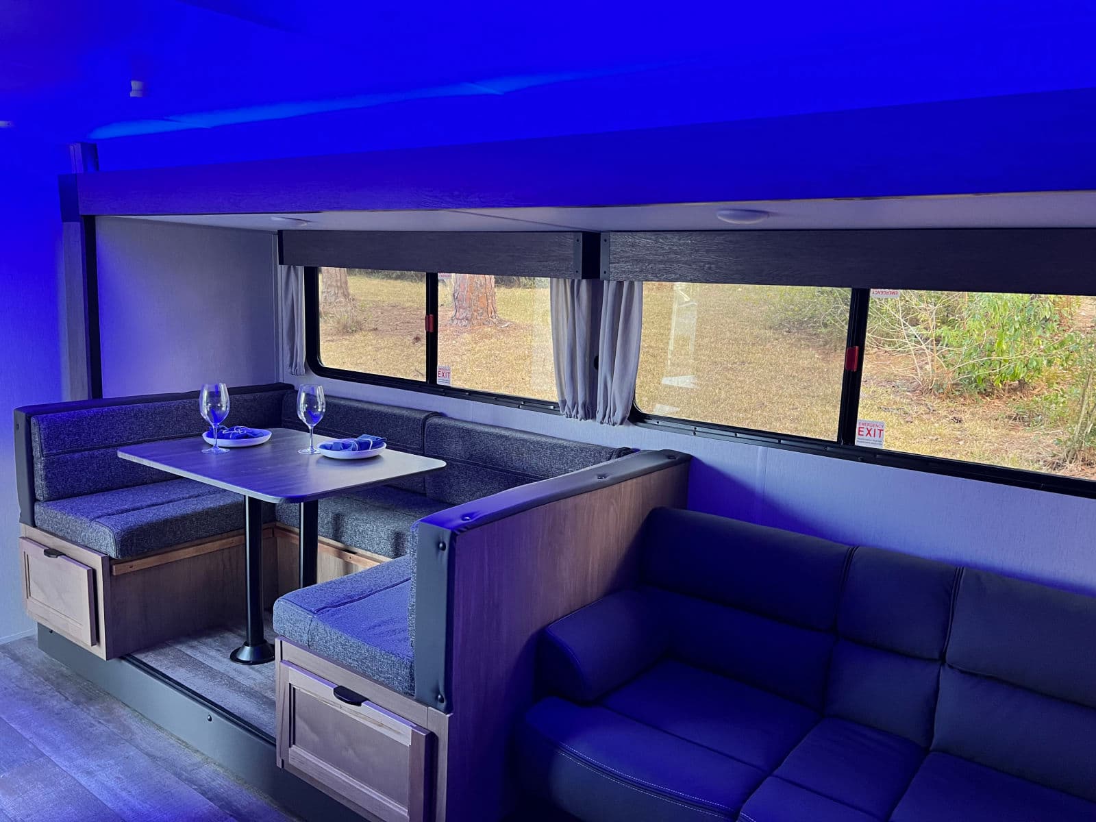 This RV rental interior features a cozy dining booth, a comfortable sofa, and windows, all under ambient blue lighting.