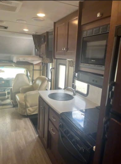 Bright RV rental interior showing a compact kitchen with sink & stove, overhead bunk bed, and cozy chairs in the living area.