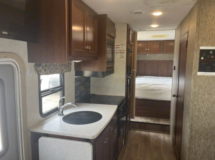 Inside this rv rental, a compact kitchen with dark wood cabinets, sink, and stove transitions to a bedroom area.