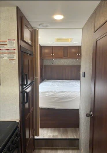 RV rental interior: A compact bedroom with a white bed and overhead cabinets, viewed from a narrow hallway with a kitchen on the left.