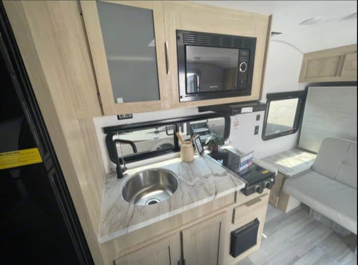 Compact RV rental kitchen features a sink, microwave, stove, and light wood cabinets, ideal for travel.