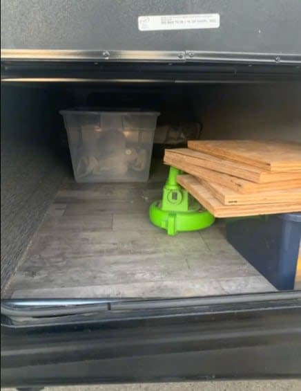 An RV rental's accessible storage compartment, showing a clear tote, green equipment, plywood, and a blue bin.