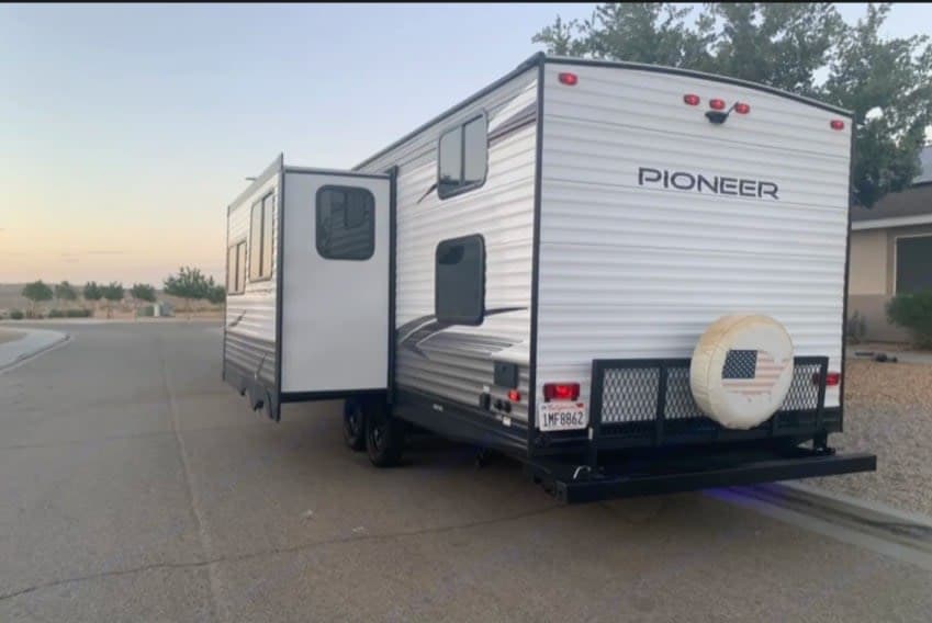 White Pioneer travel trailer RV rental with an extended slide-out, parked on a road. Note the rear cargo rack and American flag spare tire.