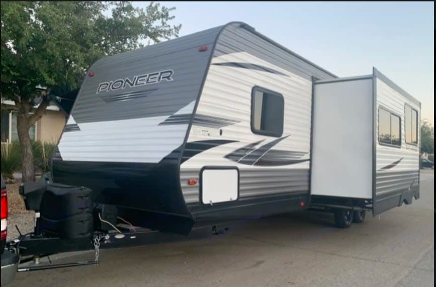 A modern gray and white Pioneer travel trailer RV rental, parked outdoors with its slide-out extended, awaits adventure.