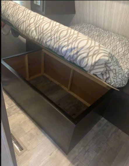RV rental bed with mattress lifted, exposing a large under-bed storage compartment. Features patterned bedding & dark frame.