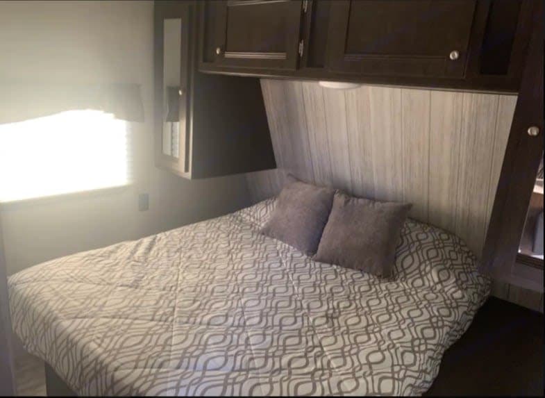 Comfortable RV rental bedroom featuring a queen bed with a patterned comforter, overhead cabinets, and a bright window.