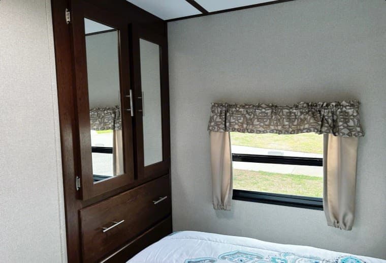 RV rental bedroom: dark wood wardrobe with mirrored doors, window with valance & curtains, and a comfy bed.
