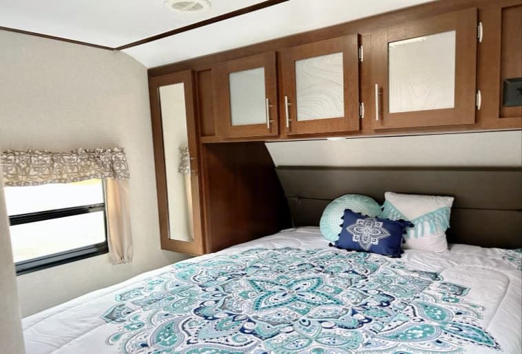 Explore this inviting RV rental bedroom, featuring a vibrant patterned bedspread, practical overhead cabinets, and a window.