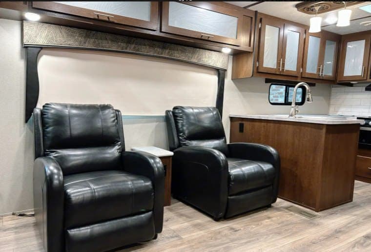 Comfortable RV rental interior featuring two black leather recliners and a modern kitchen with wooden cabinets.