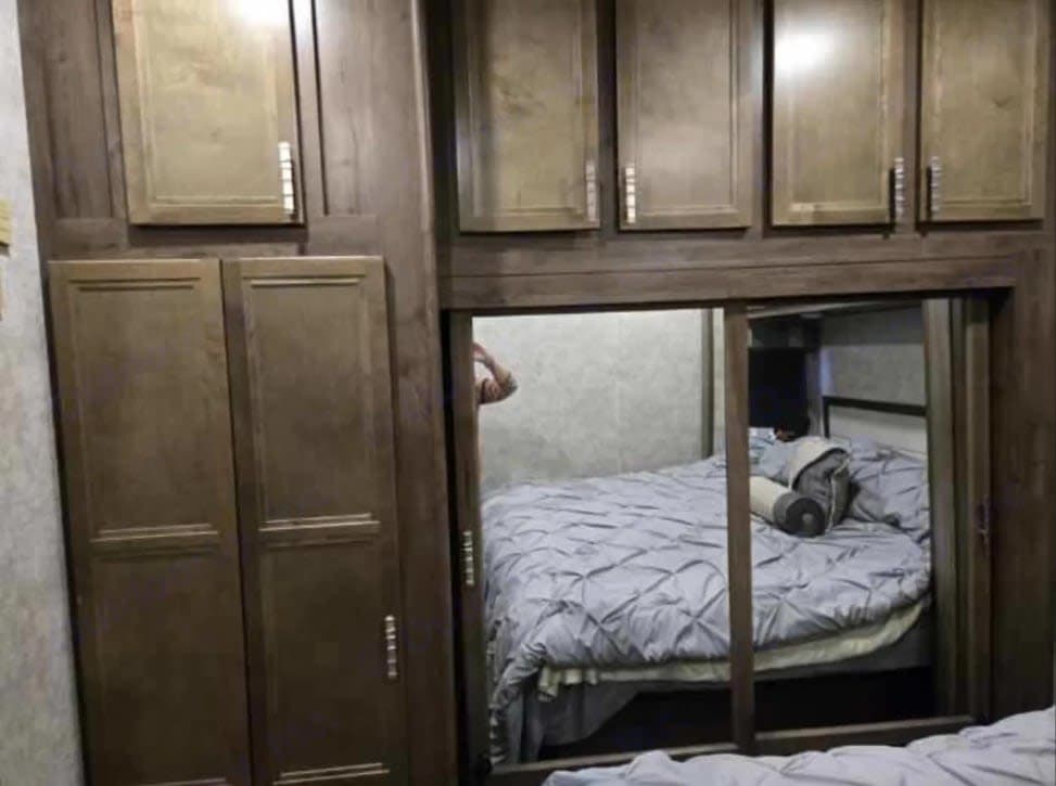 Cozy rv rental bedroom featuring wooden cabinets & a mirrored sliding door reflecting a bed with gray bedding. Person's reflection visible.