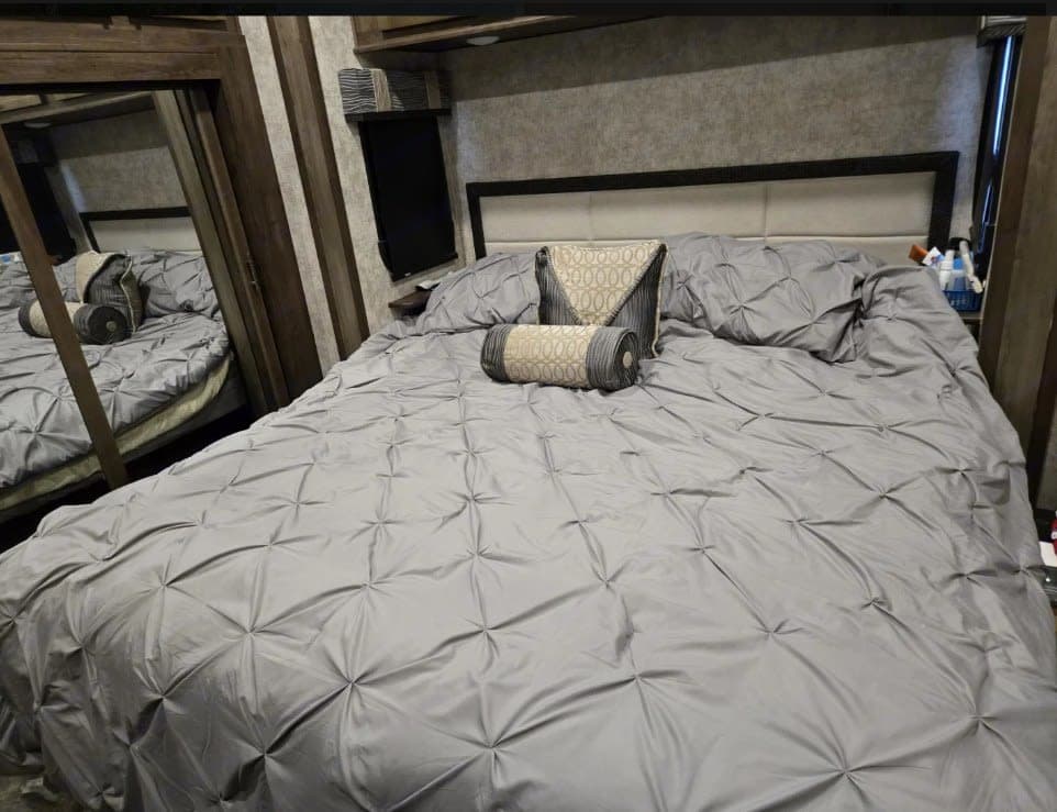 RV rental's spacious bedroom with a comfortable bed, grey pintuck bedding, and decorative pillows. Mirror on left.
