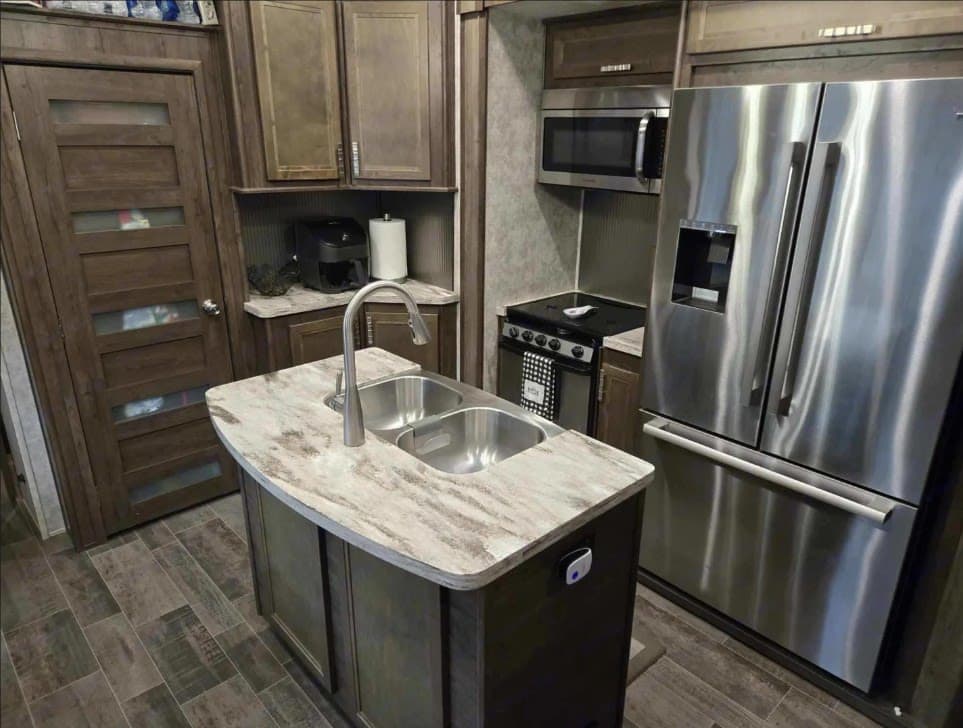 This RV rental kitchen features a modern island with double sink, stainless steel appliances, and rich wood cabinetry.