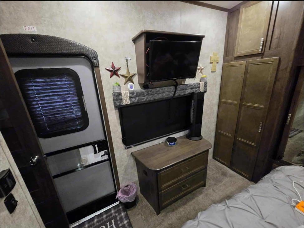 This RV rental features a cozy interior with a door, stacked TVs, a dresser, cabinets, and a bed, maximizing space.