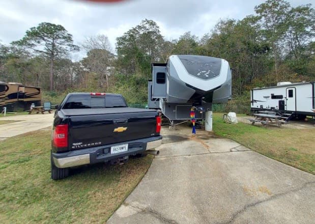 Black pickup truck parked next to a large grey fifth-wheel RV rental at a scenic campsite with other RVs under trees.