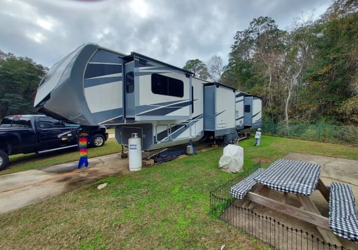 Large fifth-wheel RV rental set up at a campsite with a black truck, picnic table, and propane tanks.