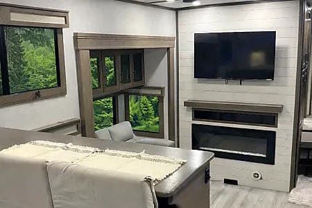 Spacious RV rental living area featuring a fireplace, TV, and scenic nature views from large windows. Ideal for travel & camping adventures.