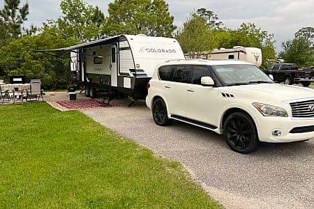White SUV towing an RV rental (Colorado travel trailer) at a campsite. Features an outdoor living area with awning and furniture.