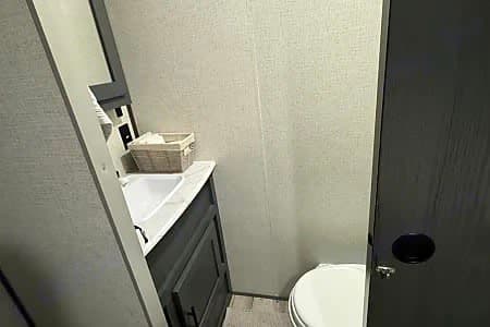 Compact RV rental bathroom. Features a vanity with a sink and storage, a mirror, and a toilet to the right. Includes a towel basket.