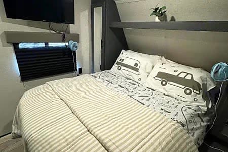 Cozy RV rental bedroom: bed with striped comforter & adventure-themed pillows. TV, window, shelf, and small fans visible.