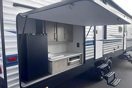 An RV rental's open side compartment reveals an outdoor kitchen: black fridge, sink, cabinets, and pull-out grill for outdoor cooking.