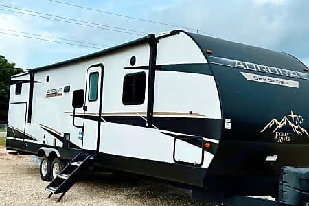 White & black Aurora Sky Series travel trailer RV rental with awning & steps, parked outdoors.