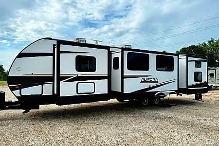 Spacious Aurora rv rental with extended slide-outs, parked on a gravel lot under a cloudy sky. Ready for adventure.