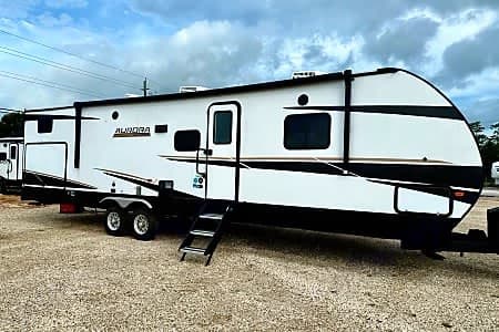 This white Aurora travel trailer, ready for your next adventure, is available for rv rental. Parked on gravel, it features black/gold accents and entrance steps.