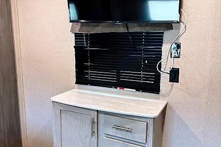 This RV rental features a wall-mounted TV over a window with blinds, and a light-colored storage cabinet below. A functional interior.