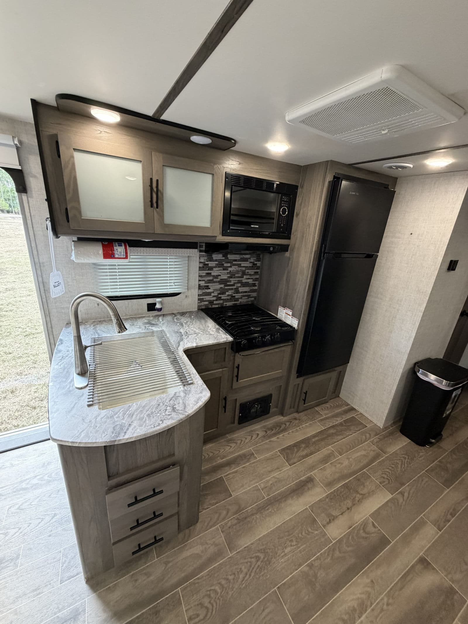 This modern RV rental kitchen features a sink, stovetop, microwave, and refrigerator, with wood-look flooring and light cabinets.