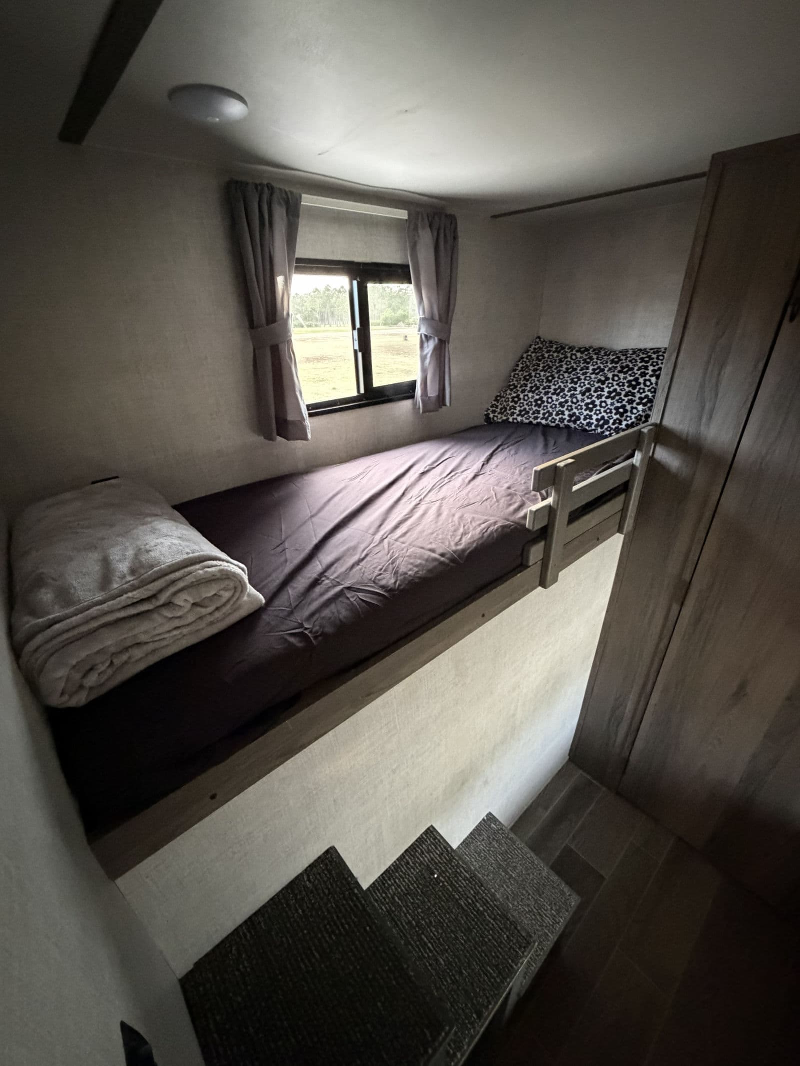 Cozy bunk bed with window and steps in an RV rental. A rolled blanket and patterned pillow add comfort to this compact space.