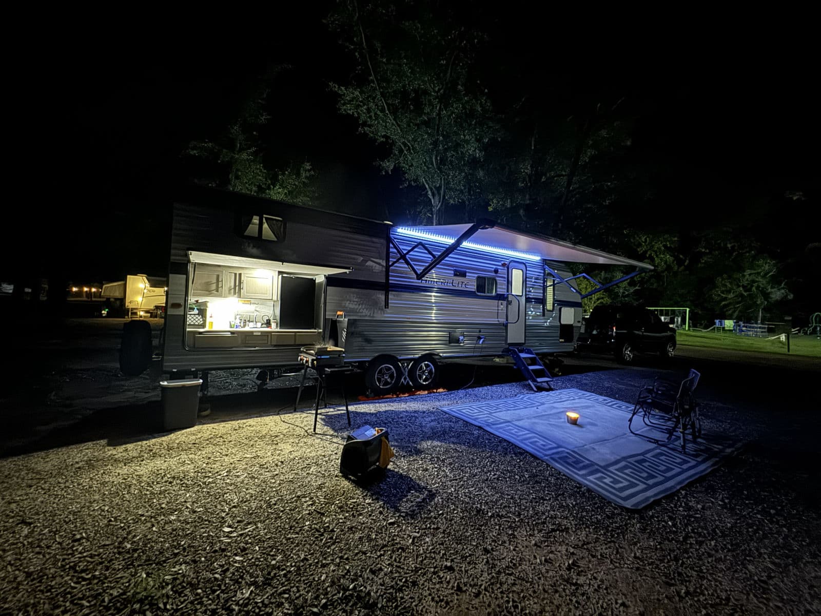 Night at an RV rental campsite: illuminated travel trailer, open outdoor kitchen, blue LED awning, and cozy patio setup.