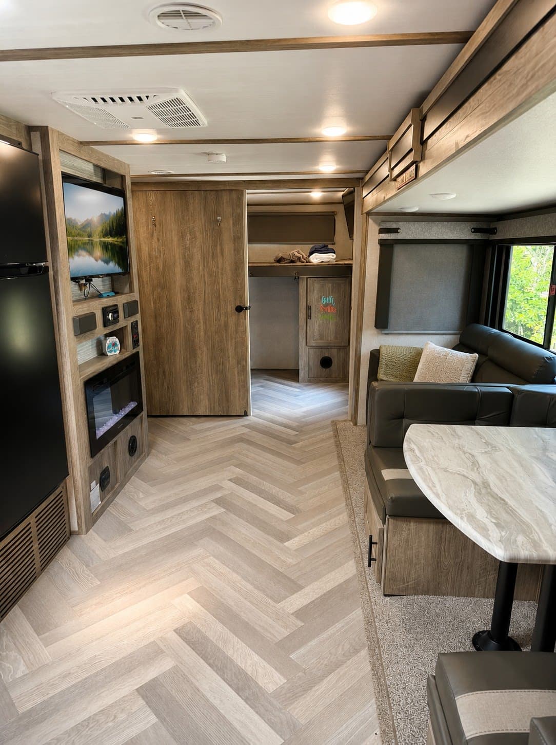 Cozy RV rental interior showcases herringbone floors, a dark sofa, dining booth, TV, and fireplace. Modern camper living space.