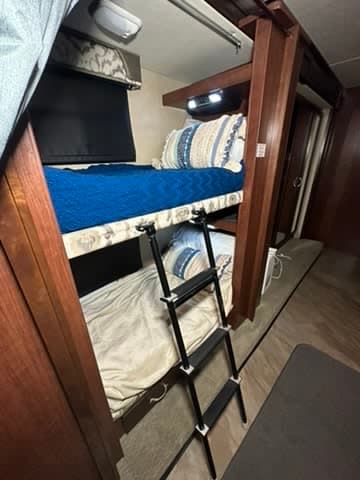 RV rental bunk beds: blue-covered top bunk, light lower bunk, and a black access ladder, all framed in wood.