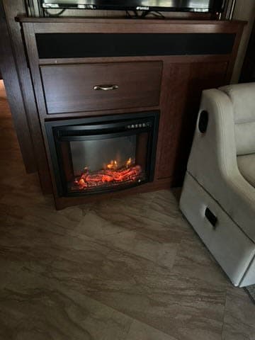 This RV rental interior features a cozy electric fireplace with artificial flames, a TV, and part of a light recliner on a patterned floor.