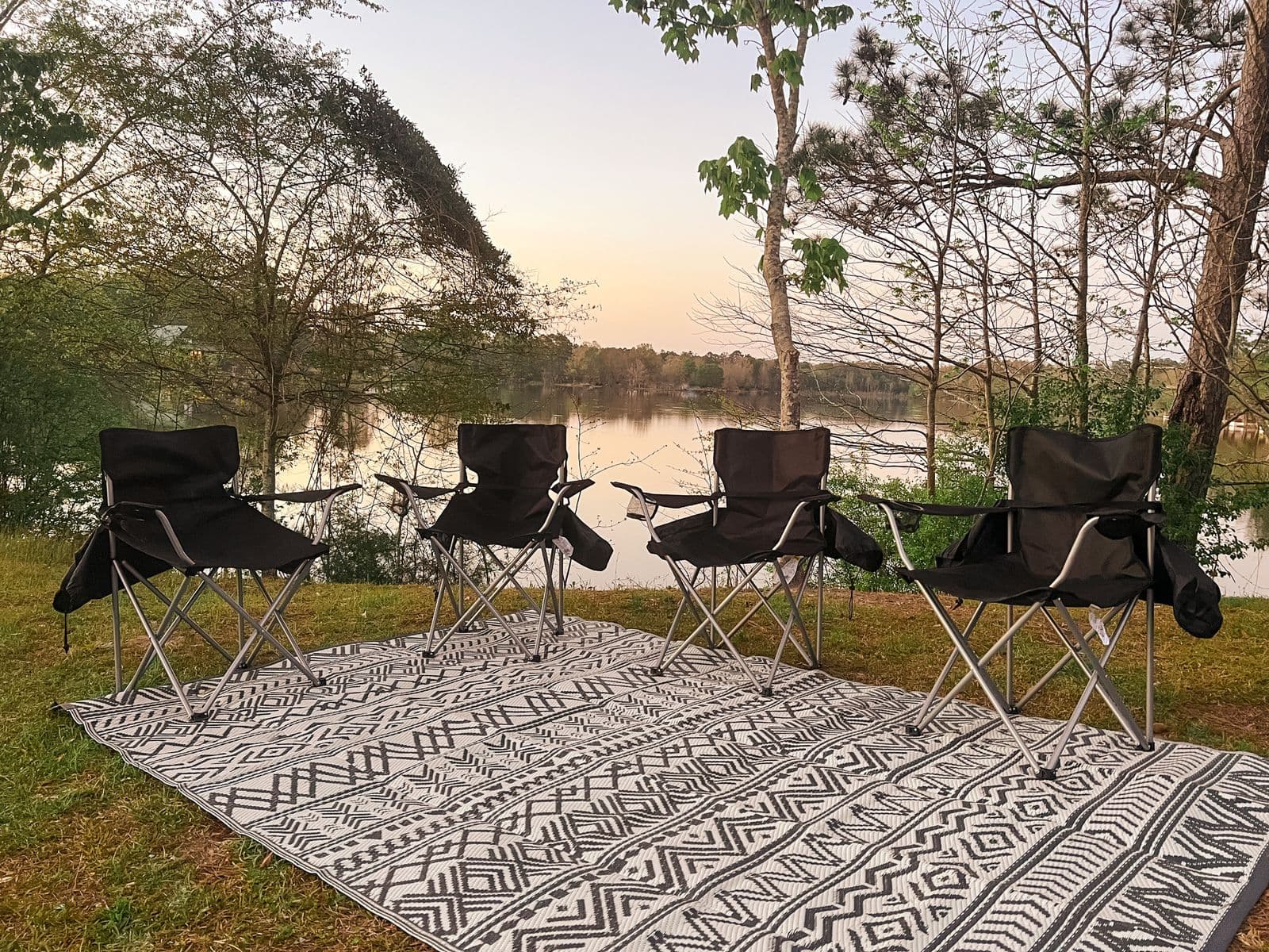 Four camping chairs on an outdoor rug beside a lake at sunset