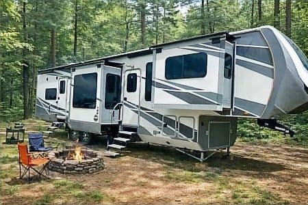A fifth wheel camper set up at a wooded campsite with chairs around a fire pit