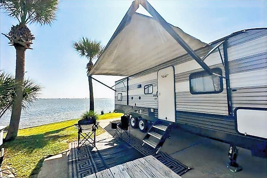 A travel trailer with its awning out on a waterfront site lined with palm trees